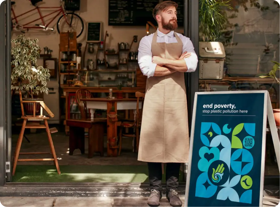 A small business owner stands proudly at the entrance of his café, wearing an apron with arms crossed. Beside him, a signboard displays a message supporting the Plastic Bank partnership: “End poverty, stop plastic pollution here,” featuring vibrant icons and a QR code.