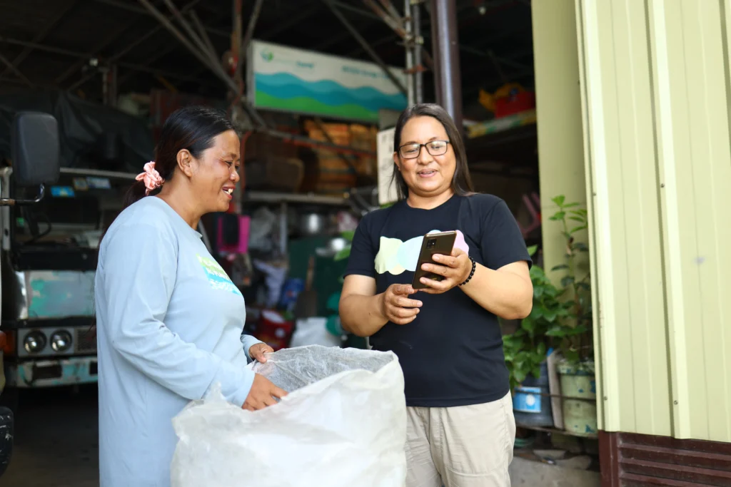 Two women smiling while using a smartphone, standing next to a large collection sack at a recycling or collection facility.