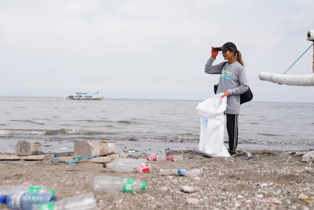 Person collecting plastic bottles and trash on a beach, holding a large white sack, with a boat visible on the ocean in the background.
