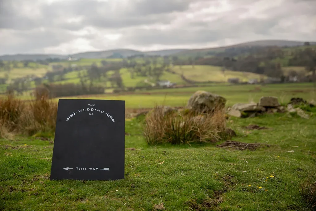 A rustic, black wedding sign on a stand in a green, grassy field. The sign has the words "THE WEDDING OF" and a small branch design at the top, along with arrows and the words "THIS WAY" at the bottom. In the background are rolling hills, a cloudy sky, and scattered rocks and foliage.