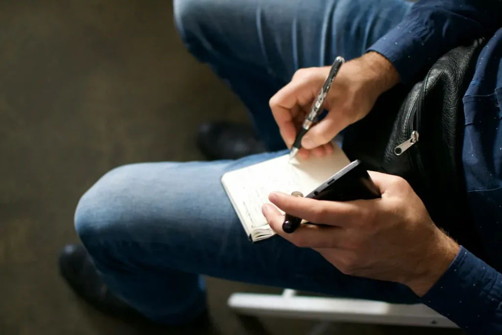 Person writing notes in a notebook while holding a smartphone, seated in casual jeans and a blue shirt.