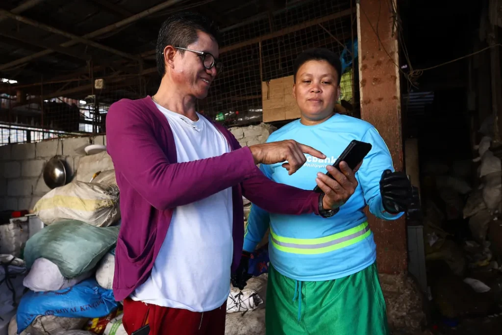Two people using a mobile phone inside a recycling facility, with sacks of sorted plastic in the background.
