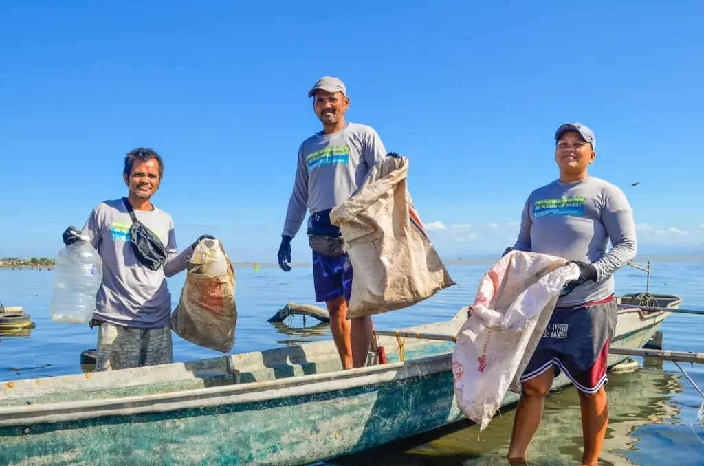 3 plastic collectors near the shore standing by a fishing boat with their plastic collection sacks